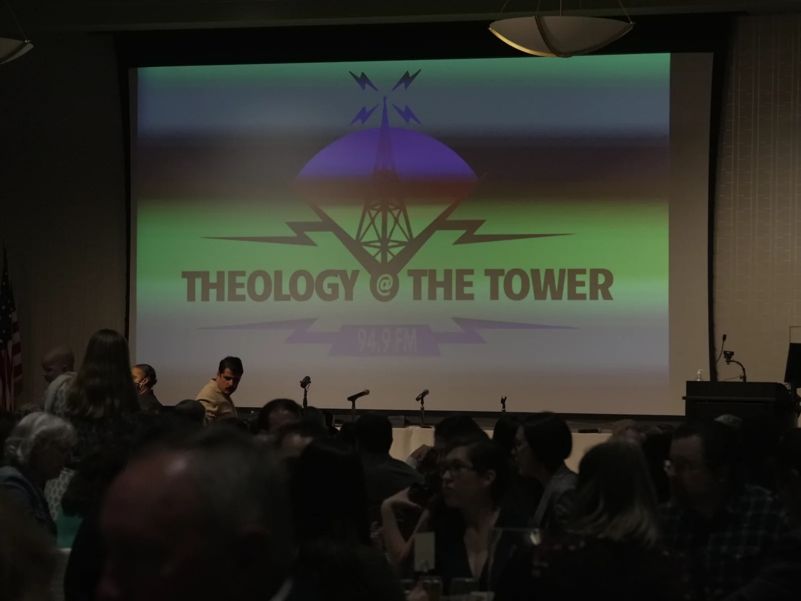 Guests seated at banquet tables face a large projection screen displaying the Theology at the Tower logo and 94.9 FM in a dimly lit event hall.
