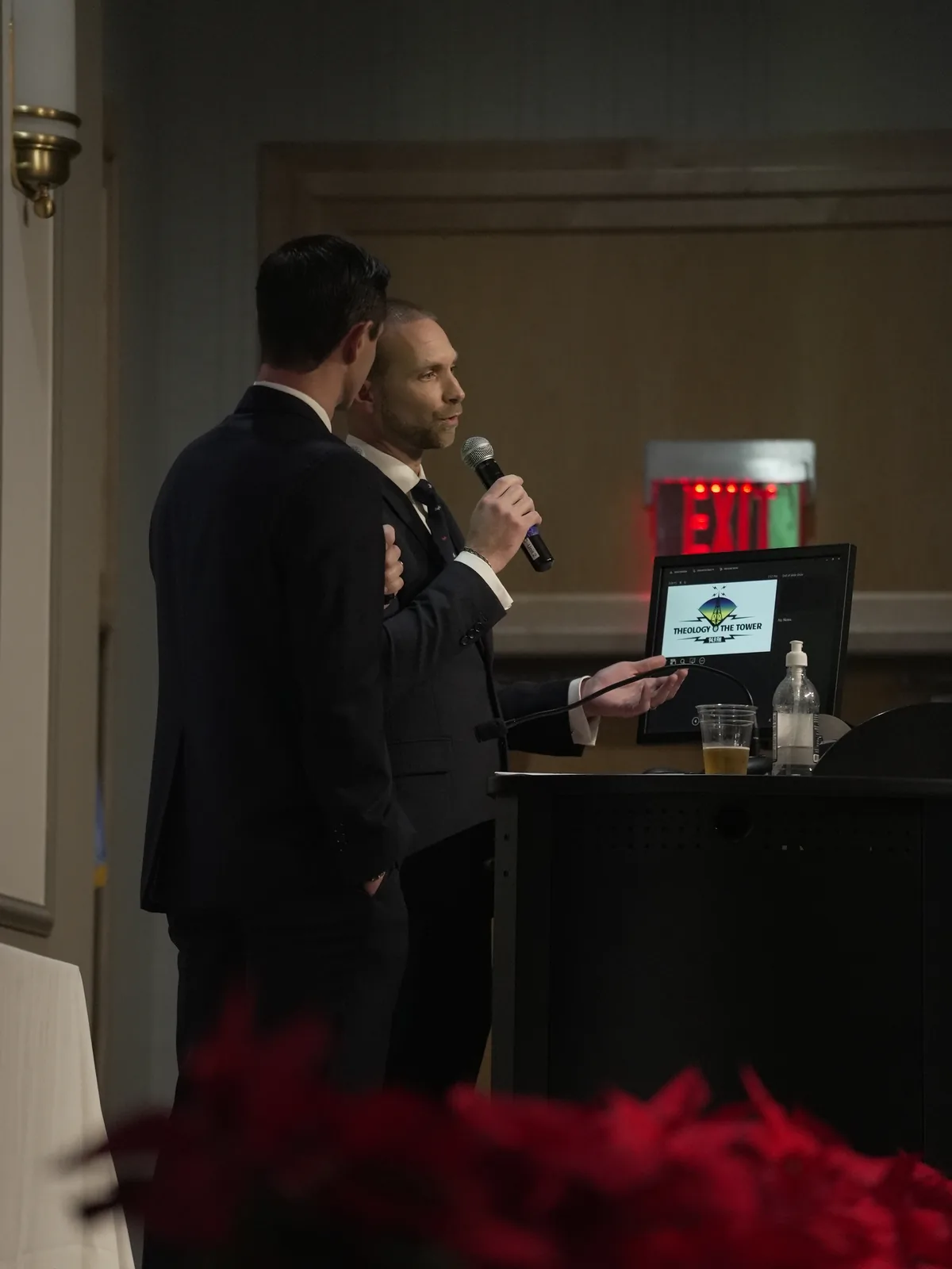 A host in a suit gestures toward a monitor showing the Theology at the Tower logo while speaking at a podium during the fundraising dinner.
