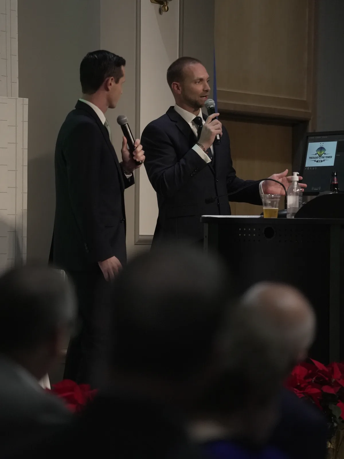 Two hosts in dark suits speak at a podium during Theology at the Tower, with the event logo on a monitor and red poinsettias in the foreground.