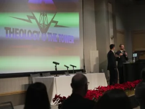 Wide view of a Theology at the Tower stage with the event logo on a projection screen, hosts at a podium, and a row of red poinsettias along the front of the stage.